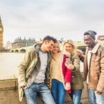 Happy multiracial friends group using smartphone in London. Mixed race millennials people lifestyle concept. Friends sharing trip on social network. Big ben and Westminster parliament on background.