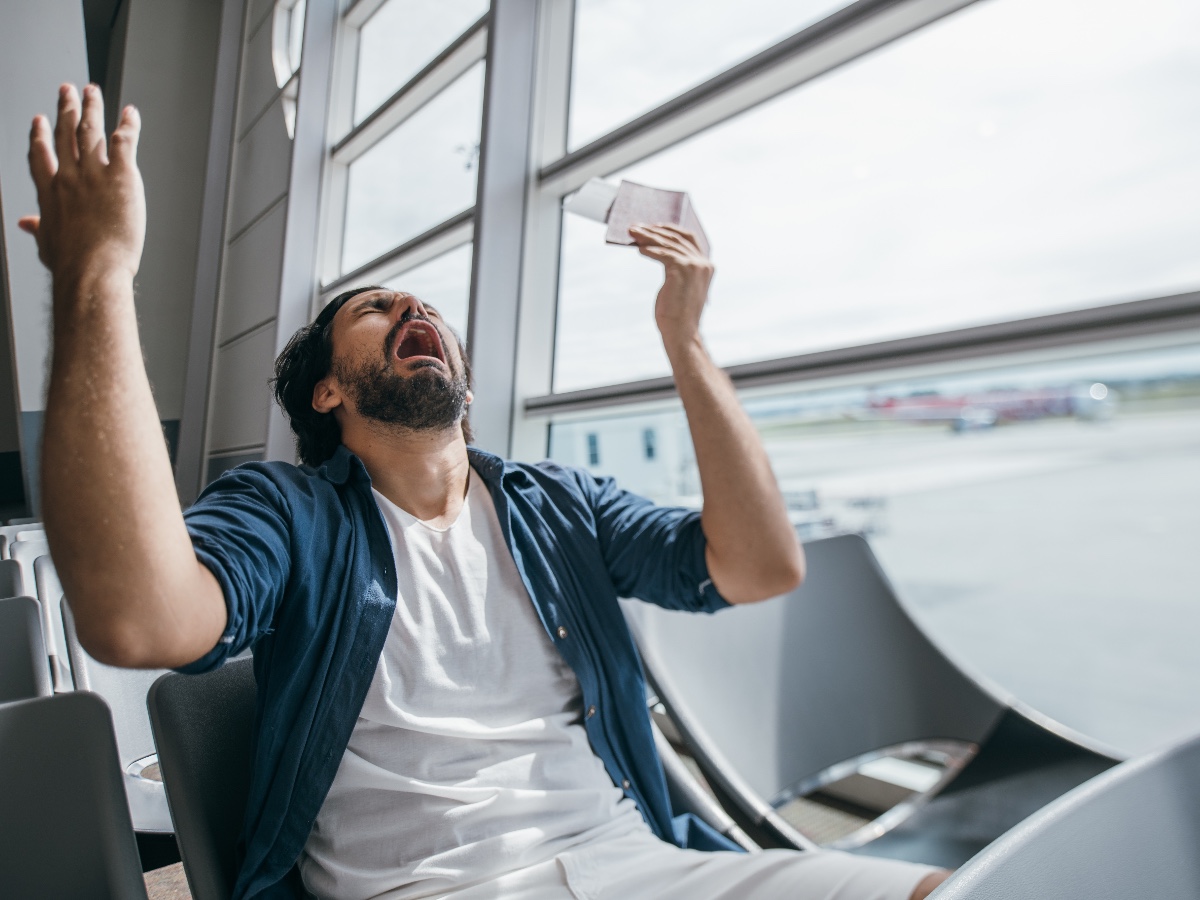 The man is very upset in the waiting room at the gate to the airport. A young guy, a traveler, waiting for a plane to fly out against the background of a large window, is very sad.