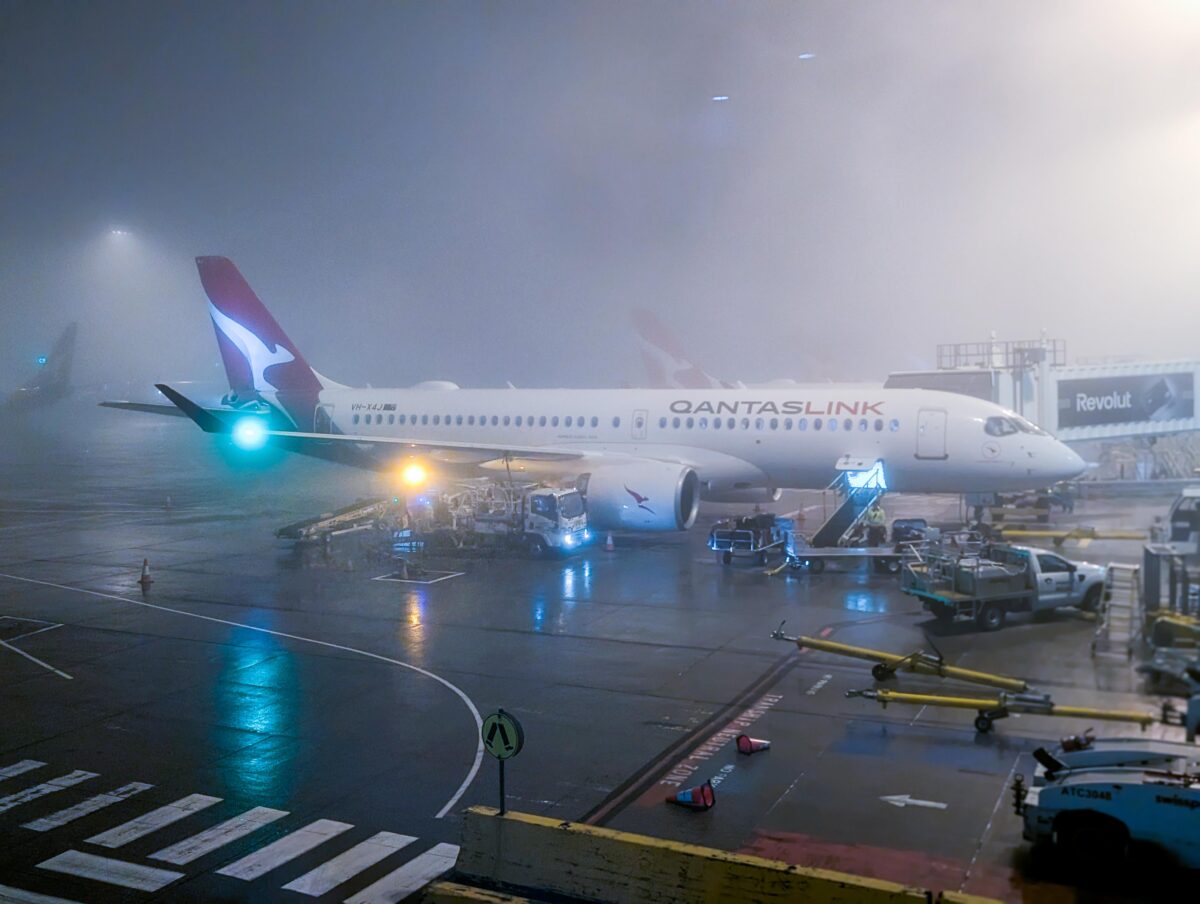 QantasLink Airbus A220 parked at a gate in thick fog at Melbourne Airport before dawn
