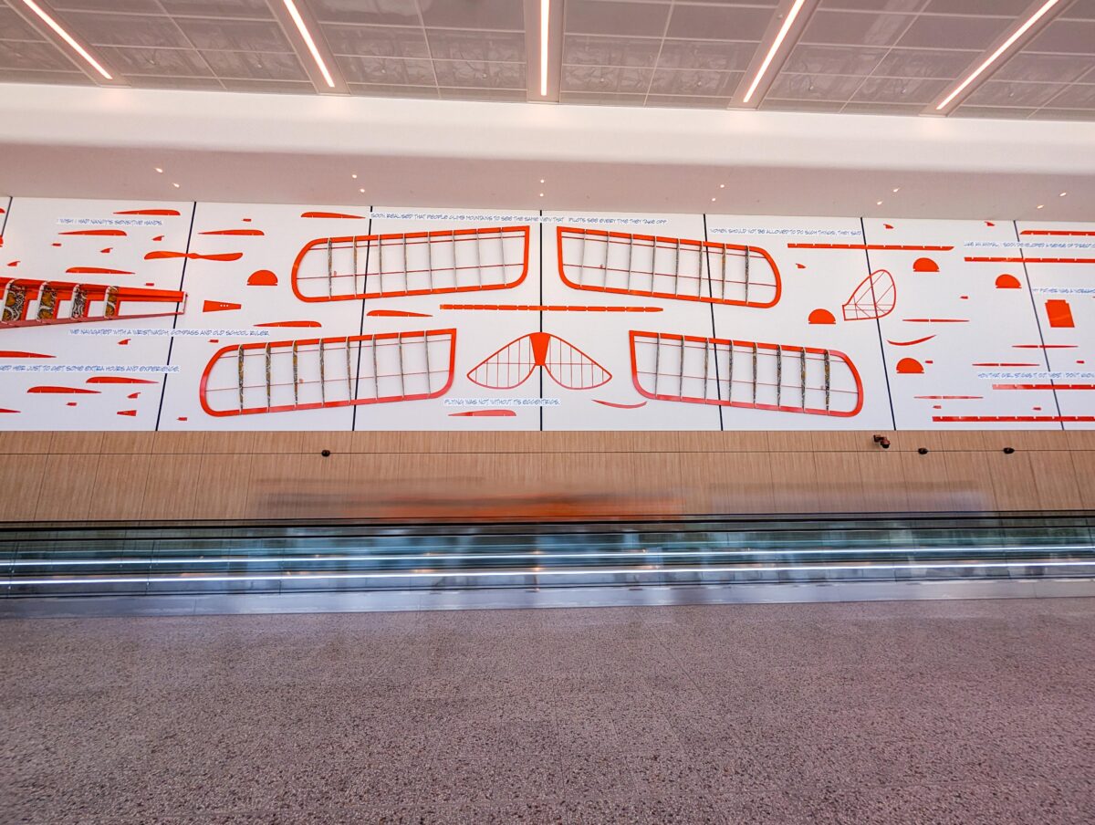 Orange aircraft wing-frame artwork with lines of text above a moving walkway at WSI